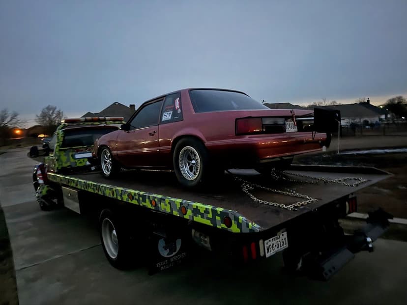 Red sports car being towed on a pickup truck with a camouflage design at dusk.