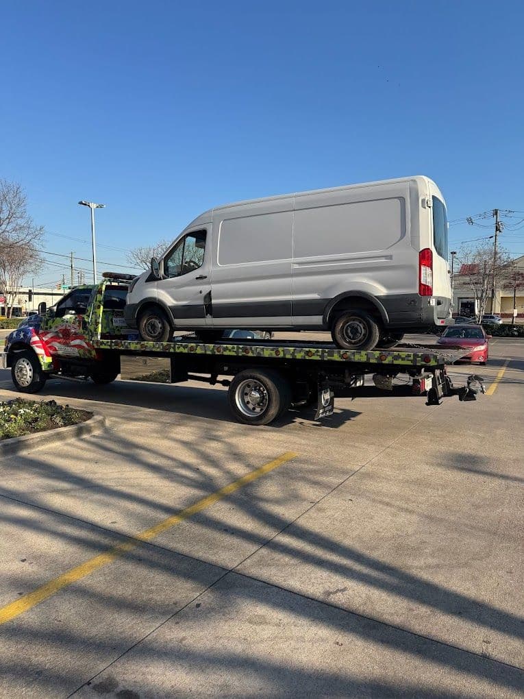 Tow truck transporting a silver van under a clear blue sky in a parking lot.
